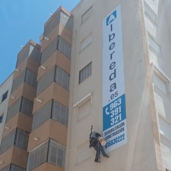 Obrero colgado con arneses en la fachada de un edificio junto con un cartel de Albereda con los teléfonos